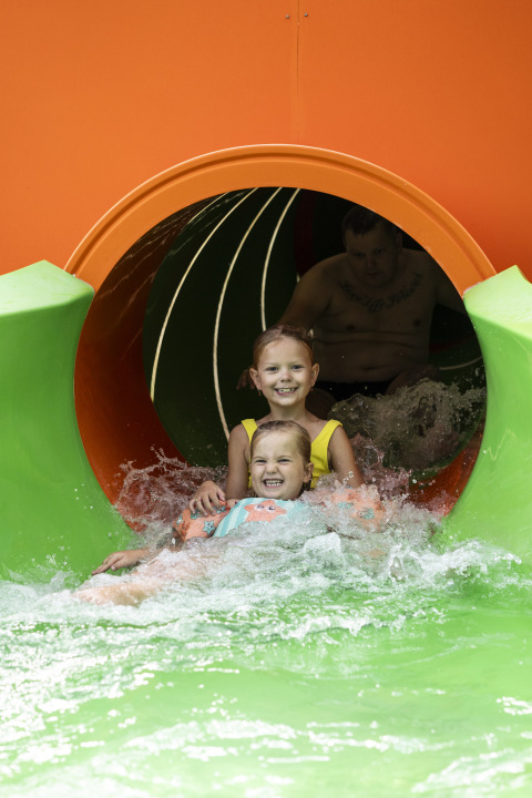 Dos niños sonrientes y un adulto salen de un colorido tobogán acuático en un parque acuático con agua salpicando.