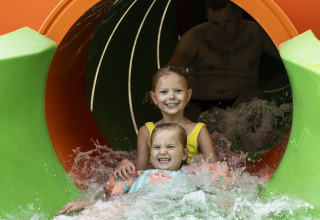 Dos niños sonrientes y un adulto salen de un colorido tobogán acuático en un parque acuático con agua salpicando.