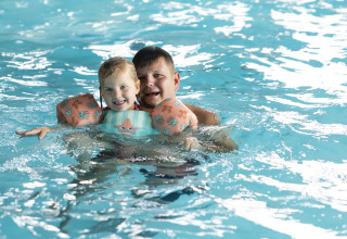 Ein Kind mit Schwimmflügeln und ein Erwachsener schwimmen gemeinsam in einem Hallenbad.