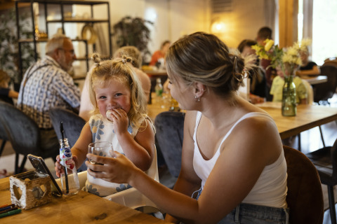 Una niña y una mujer sonríen juntas mientras se sientan en una mesa de madera en una cafetería glamping.