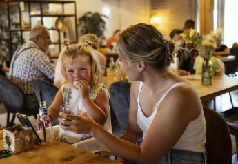 A young girl and a woman enjoy drinks and laughter together at a wooden table in a glamping lounge.