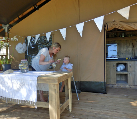 Mother feeding child in high chair inside a safari tent with kitchen and wooden table outside.