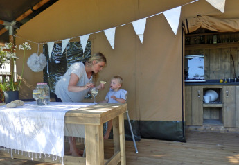 Mother feeding child in high chair inside a safari tent with kitchen and wooden table outside.