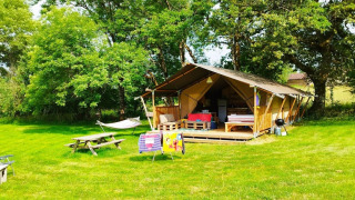 Een gezellige safaritent met veranda, hangmat, picknicktafel en stoelen op een grasveld met bomen rondom.