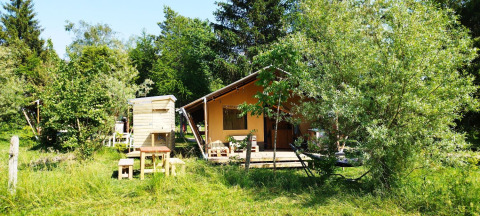 Safari tent with bathroom among lush green trees and grass, wooden outdoor furniture in front.