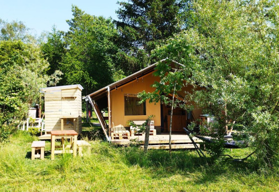 Tente safari avec salle de bain entourée d’arbres et d’herbe, mobilier extérieur devant.