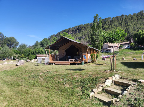 Tienda safari llamada Lodgetent met badkamer en un campo verde con árboles y colinas bajo un cielo azul.