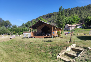 Tienda safari llamada Lodgetent met badkamer en un campo verde con árboles y colinas bajo un cielo azul.