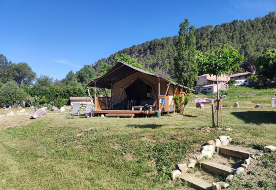 Tienda safari llamada Lodgetent met badkamer en un campo verde con árboles y colinas bajo un cielo azul.