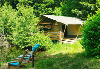 Safari tent by a lakeside surrounded by lush greenery, two chairs with blue cushions sit by the water.