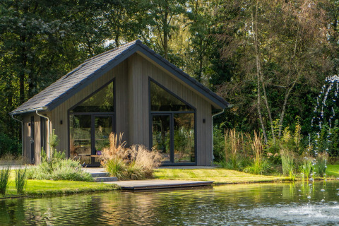 Ecovilla Waterzicht Comfort lodge by a pond, set in a natural, green landscape with large windows and trees.