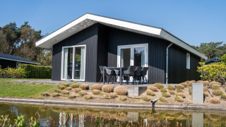 Modern black Villa Waterzicht Comfort lodge with outdoor seating by a pond, surrounded by greenery