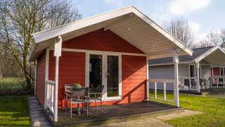 Red wooden Tiny House Deluxe with porch and chairs on a grassy lawn, neighboring tiny houses in the background.