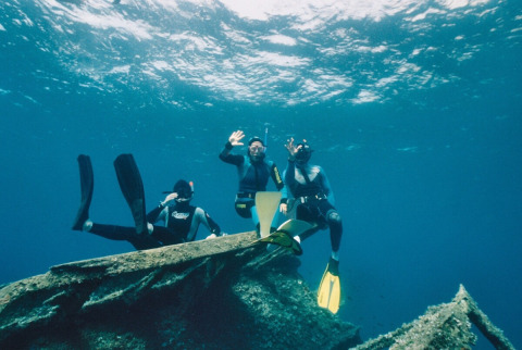 Three snorkelers explore underwater near Portoferraio, Tuscany, Italy, waving at the camera in clear blue water.