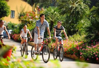 Four people enjoy cycling together along a colorful flower path at Rosselba le Palme campsite in Tuscany, Italy.