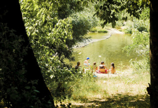 A group relaxing by a river surrounded by trees at Campsite La Plage holiday park in Occitanie, France.