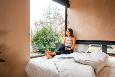 Vrouw werkt op laptop en kijkt uit het raam vanuit bed in Slowcabin-lodge te Zomerlicht, Nederland.