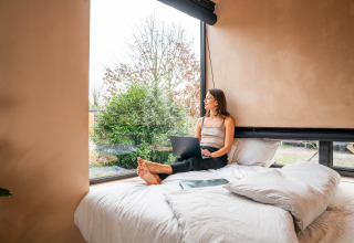 Vrouw werkt op laptop en kijkt uit het raam vanuit bed in Slowcabin-lodge te Zomerlicht, Nederland.