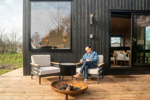 Man leest buiten aan een moderne zwarte Slowcabin-lodge met vuurkorf, terraszitjes en groot raam.