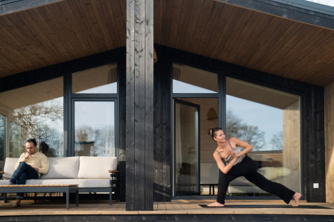 Een man leest op een bank terwijl een vrouw yoga doet op het terras bij Slowlodge Sage in Nederland.