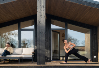 Een man leest op een bank terwijl een vrouw yoga doet op het terras bij Slowlodge Sage in Nederland.