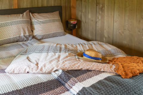 Gemütliches Bett in einer Glamping-Unterkunft mit gemusterten Decken, Strohhut und orangefarbenem Tuch.