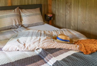 Gemütliches Bett in einer Glamping-Unterkunft mit gemusterten Decken, Strohhut und orangefarbenem Tuch.