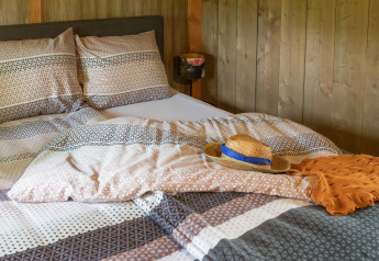 Lit douillet dans un hébergement glamping avec housses à motifs, chapeau de paille et foulard orange.