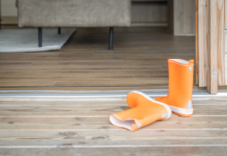 Ein Paar orangefarbene Gummistiefel steht auf der Holzterrasse am Eingang einer Glamping-Unterkunft.