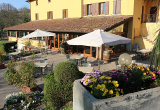 Udendørs terrasse med caféborde, parasoller og blomster foran et charmerende gul landhus i Lombardiet, Italien.