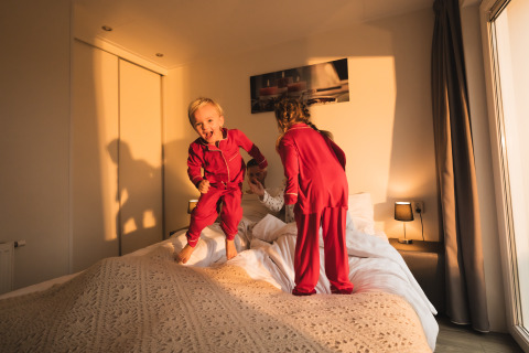 Twee kinderen in rode pyjama’s springen vrolijk op bed in een zonnige kamer in Resort Mooi Bemelen.