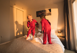 Twee kinderen in rode pyjama’s springen vrolijk op bed in een zonnige kamer in Resort Mooi Bemelen.