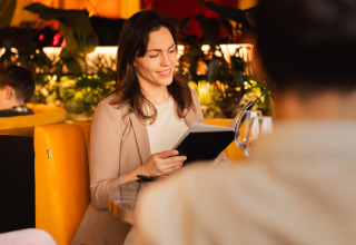 Vrouw glimlacht met een notitieboek op een gele zitbank in een lodge bij Schaelsbergerbosch, Resort Mooi Bemelen.