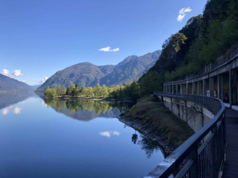 Vista sul lago tranquillo al Campeggio Belvedere in Lombardia, Italia, con montagne e alberi sotto il cielo sereno.