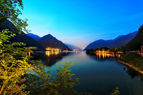 Vista serale su lago e montagne dal campeggio Belvedere in Lombardia, Italia, con luci riflesse sulla riva.
