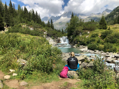 Persona seduta vicino al fiume con zaino al Campsite Belvedere, circondata da montagne in Lombardia.