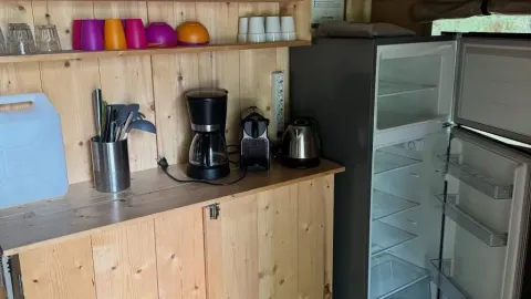 Et lille køkkenområde i et safaritelt med køleskab, kaffemaskine, elkedel og farvede kopper på hylderne.