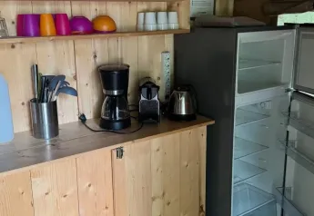 Et lille køkkenområde i et safaritelt med køleskab, kaffemaskine, elkedel og farvede kopper på hylderne.