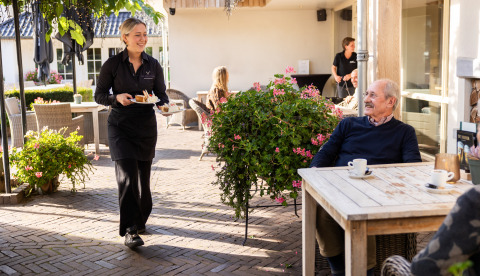 Une serveuse apporte un dessert à un homme âgé sur la terrasse extérieure d’un hébergement glamping.