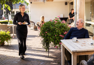 Une serveuse apporte un dessert à un homme âgé sur la terrasse extérieure d’un hébergement glamping.
