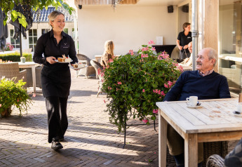 Une serveuse apporte un dessert à un homme âgé sur la terrasse extérieure d’un hébergement glamping.