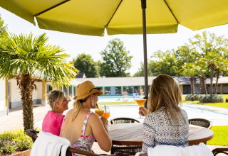Trois femmes dégustent des cocktails à une table extérieure sous un parasol près d'une piscine et tiny house.