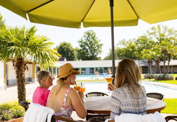 Trois femmes dégustent des cocktails à une table extérieure sous un parasol près d'une piscine et tiny house.