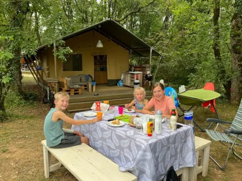 Famille prenant un repas à une table de pique-nique devant une tente safari lors d’un séjour en forêt.