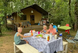 Famille prenant un repas à une table de pique-nique devant une tente safari lors d’un séjour en forêt.