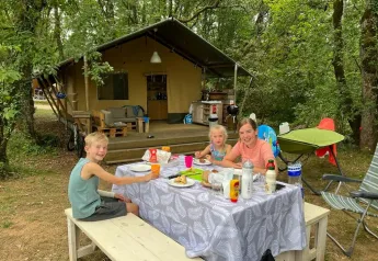 Family enjoying a meal at a picnic table in front of a safari tent on a wooded camping trip.