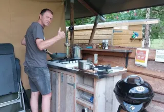 Uomo cucina in cucina all'aperto vicino a una tenda safari, pollice in su e sorriso rilassato.