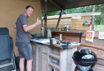 Homme cuisine dans une cuisine extérieure près d'une tente safari, pouce levé et sourire détendu.
