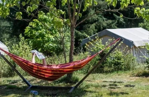 Une personne se détend dans un hamac avec un journal près d'une tente safari entourée de verdure.