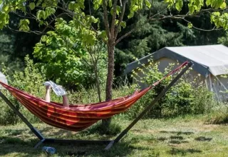 A person relaxes in a hammock reading a newspaper near a safari tent surrounded by lush greenery.
